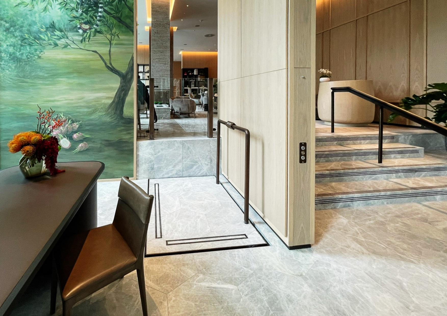 Modern interior with a compact platform lift positioned beside a short staircase, featuring light wood paneling, marble flooring, handrails, and a painted mural on the adjacent wall.