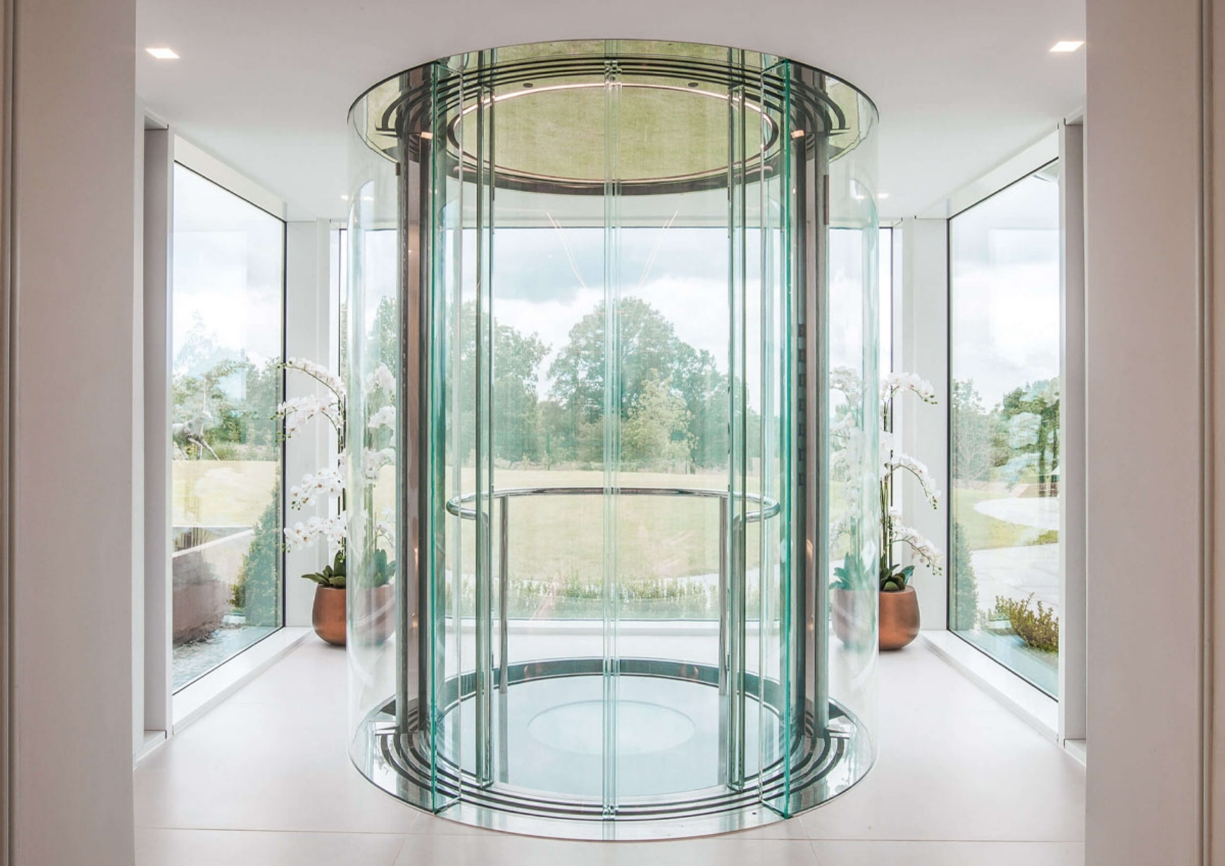 Circular glass lift in a bright modern interior, surrounded by floor‑to‑ceiling windows with views of a landscaped garden and white orchid plants on either side.